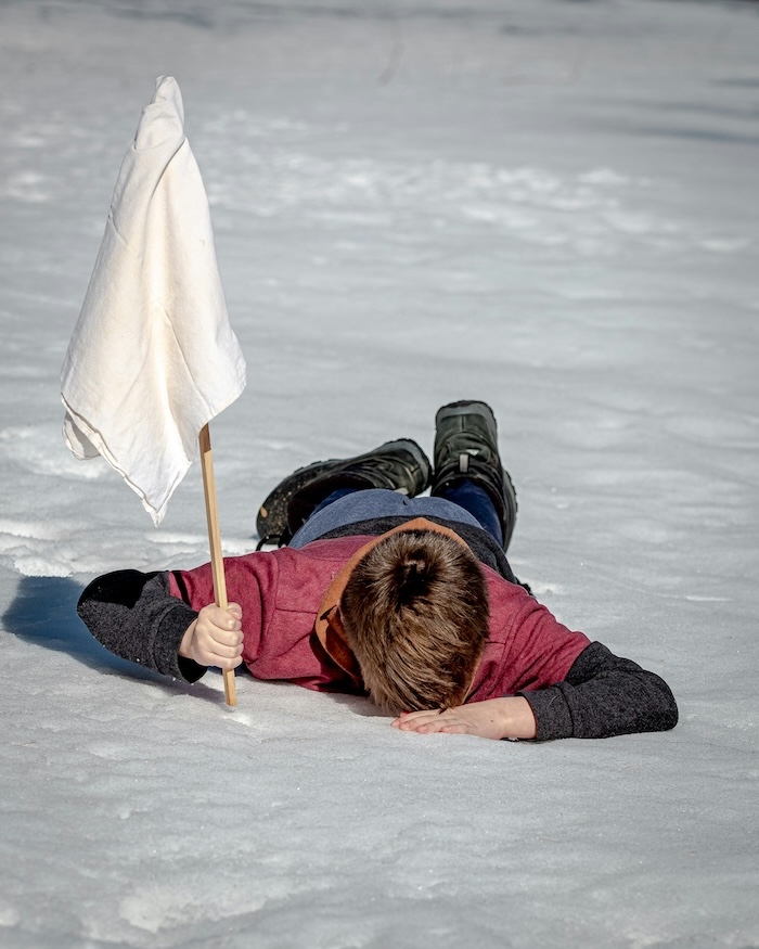Boy laying face down on snow holding up a white flag